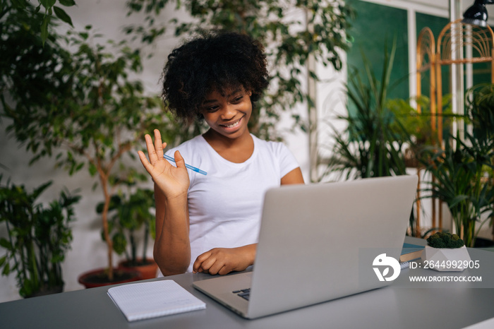 Friendly African-American female student waving hand hello at webcam of laptop, speaking on video conference talking to teacher in light home office room with modern biophilic interior.