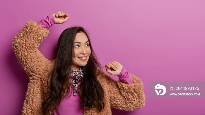 Joyful Korean woman makes victory dance, enjoys having awesome day, turns gaze aside, smiles broadly and moves with rhythm of music, dressed in stylish clothes, isolated on lilac background.