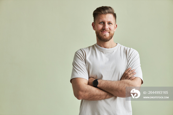 Waist up portrait of bearded young man smiling at camera while standing with arms crossed against pastel green, minimal