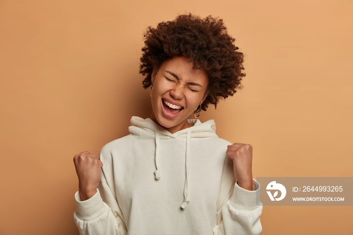 Photo of overemotive curly ethnic woman rejoices praises or winning competition, makes fist pump with closed eyes, tilts head and smiles happily, stands in triumph pose, wears casual white hoodie