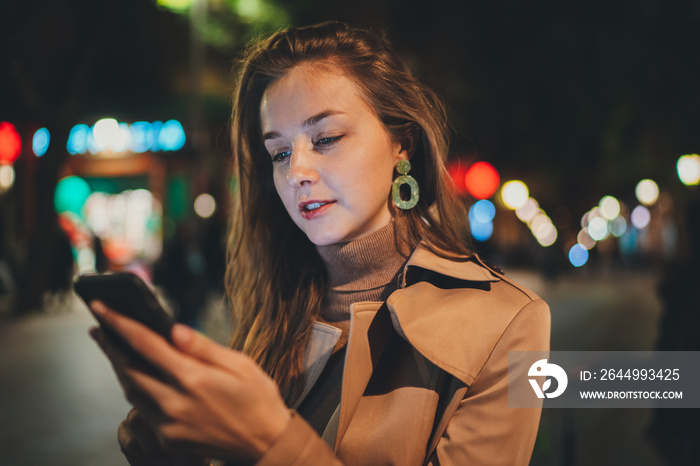 Young beautiful woman using cellphone sharing post in social networks during night walk in the city, Attractive girl reading sms message or browsing internet searching free wifi connection outdoors