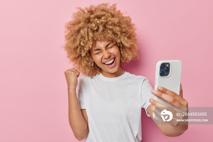 Overjoyed lucky European woman with curly bushy hair does winner gesture clenches fist takes selfie on smartphone dressed in casual white t shirt isolated over pink background uses filter app