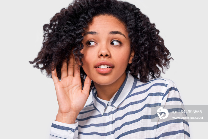 Curious African American young woman paying attention and placing hand on ear asking someone to speak louder or whisper, isolated on white wall. Pretty Afro girl which overhears secret conversation.