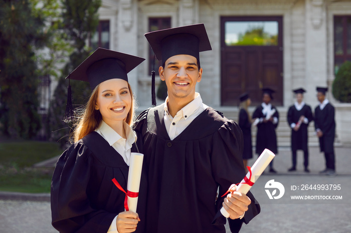 Portrait of happy graduates. Two friends in graduation caps and gowns standing outside university building with other students in background, holding diploma scrolls, looking at camera and smiling