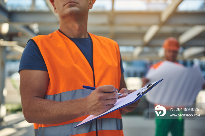 Cropped photo of adult builder in orange vest writing documents while his male colleague looking draft