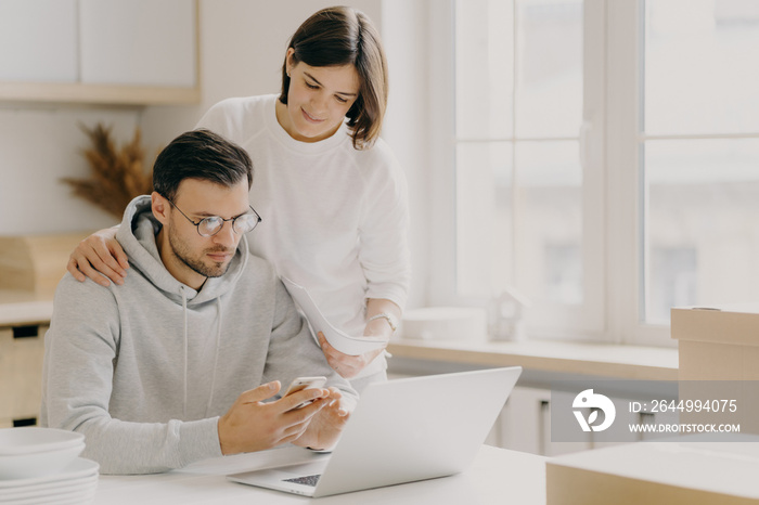 Picture of husband and wife study notification from bank, holds mobile phone and papers, work on laptop computer, pose in kitchen during relocation day, dressed in casual wear, save family money