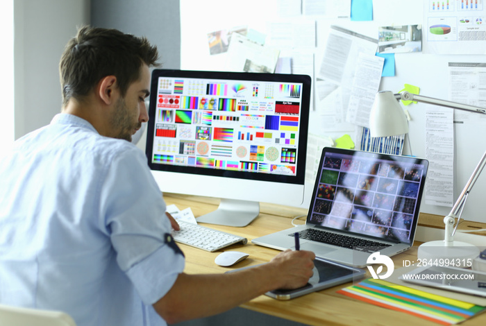 Portrait of young designer sitting at graphic studio in front of laptop and computer while working online.