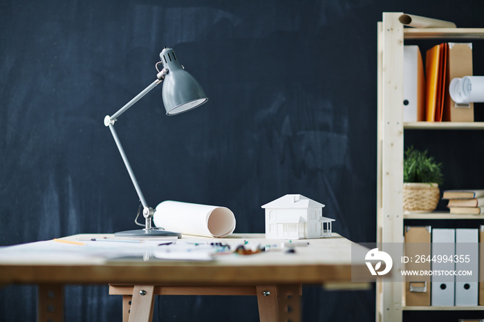 Background shot of architects workplace in modern office with house model on desk, metal lamp, blueprints and supplies against blackboard