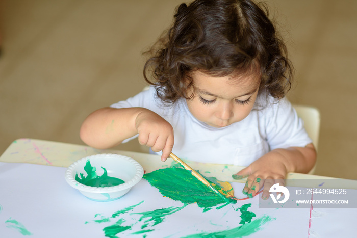 Summer or Autumn.Cute little girl drawing with green paint on fall leaves. Curly adorable child painting at table.