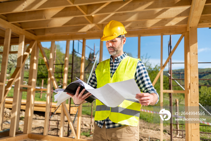 Construction engineer or architect with blueprints visiting building site of wood frame house