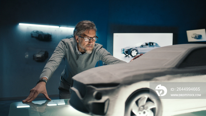 Automotive engineer touches the model of the prototype car made of clay to make final changes in the design. Grey haired car developer with framed glasses in a car manufacturing company.