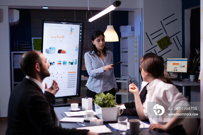 Workaholic businesswoman pointing financial strategy using monitor working overtime in company meeting office room. Diverse multi-ethnic teamwork overworked solving management statistics in evening