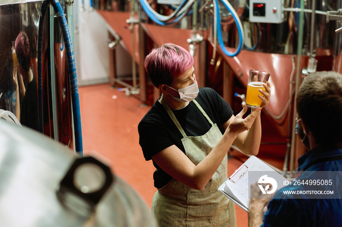 Two young contemporary brewers examining glass of fresh beer after preparation