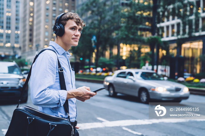 Successful male entrepreneur in formal wear holding modern smartphone device and using music application for listening audio via headphones for noise cancellation, Caucasian man in downtown