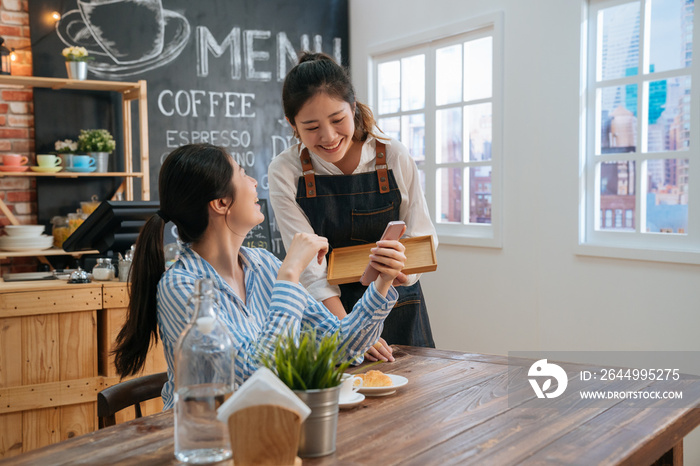 Smiling friendly waitress serving coffee drinks and croissant to friend at cafe table. cafeteria server and regular female visitors laughing together at funny joke on cellphone in modern coffeehouse.