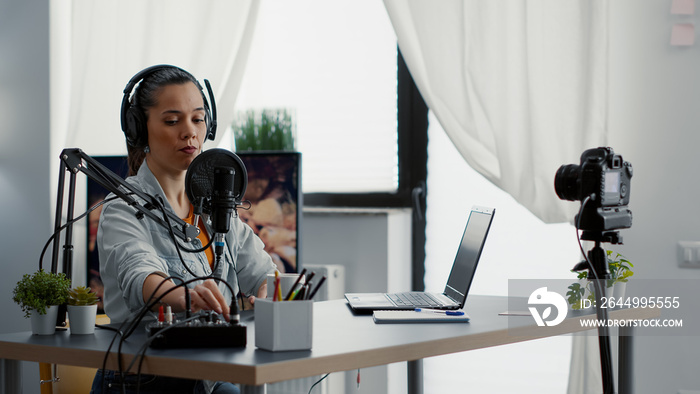 Creative content creator recording vlog in home studio while tweaking knobs on audio mixer. Internet live broadcast talk show host talking with audience while using professional audio equipment.