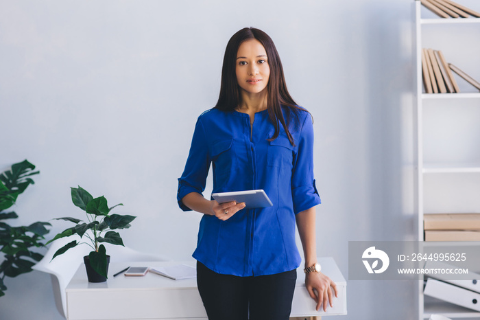 modern attractive woman holding digital tablet, looking at camera while standing at modern office.