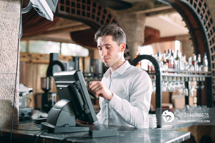 small business, people and service concept - happy man or waiter in apron at counter with cashbox working at bar or coffee shop.