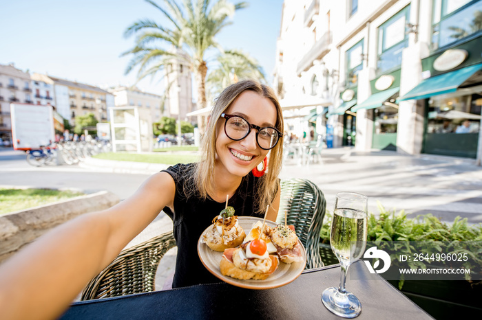 Young woman making selfie photo with pinchos, traditional spanish snack, and glass of wine sitting outdoors at the bar in Valencia city