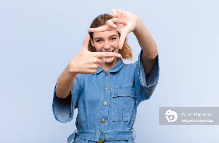 young blonde woman feeling happy, friendly and positive, smiling and making a portrait or photo frame with hands against flat color wall