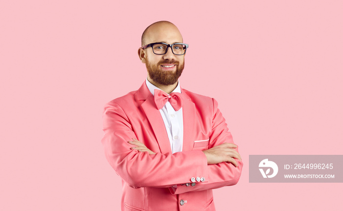 Portrait of smiling young bearded Caucasian male entertainer or performer in suit isolated on pink studio background. Happy man in glasses in party jacket stand with arms crossed.