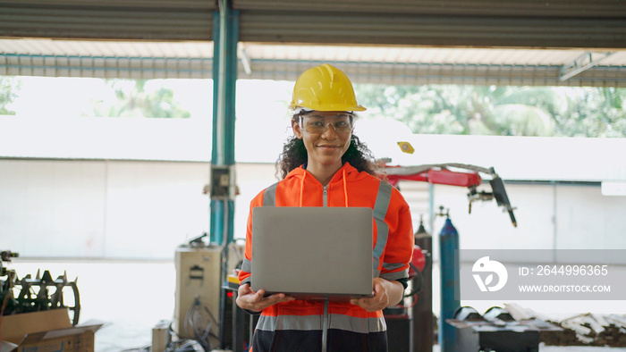 Young Africa American engineer wearing safety helmet and glasses holding the laptop and walking and smile in the robotic arm factory. Future technology and artificial intelligent concept