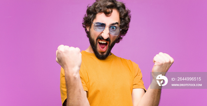 young crazy cool man shouting aggressively with an angry expression or with fists clenched celebrating success against flat wall