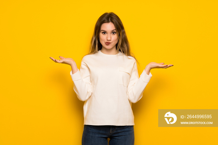 Teenager girl over yellow wall having doubts while raising hands and shoulders