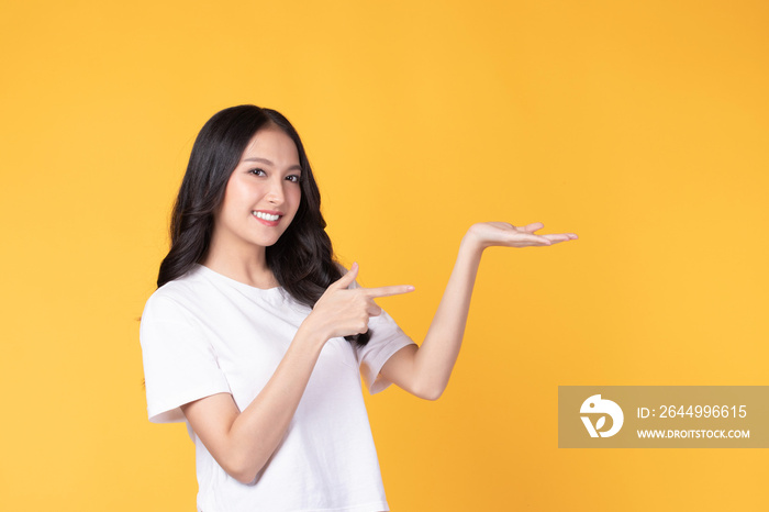 Beautiful Asian woman wearing casual clothes smile and pointing finger to an empty palm isolated on yellow background studio portrait.