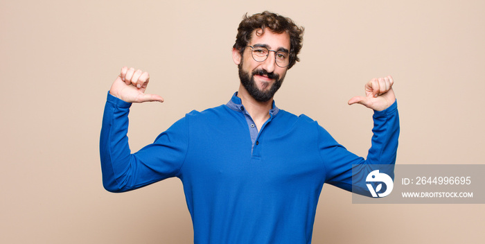 young handsome man feeling proud, arrogant and confident, looking satisfied and successful, pointing to self against flat wall