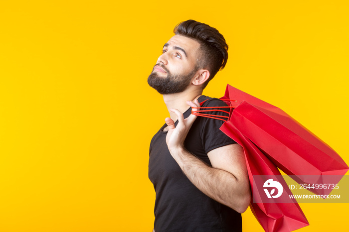 Side view of an upset young bearded stylish hipster man holding shopping bags posing on a yellow background. Concept of superfluous purchases.