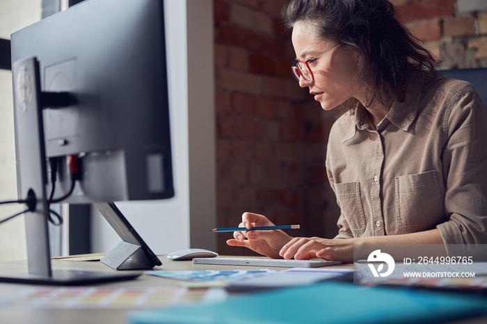 Side view of creative woman, interior architect or designer wearing glasses working on a project, using computer while sitting at the desk in her office