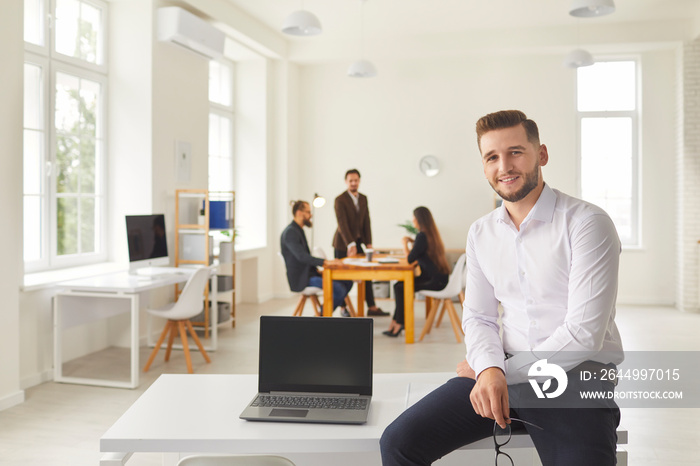 Young businessman with laptop with blank screen sitting on desk in coworking space looking at camera