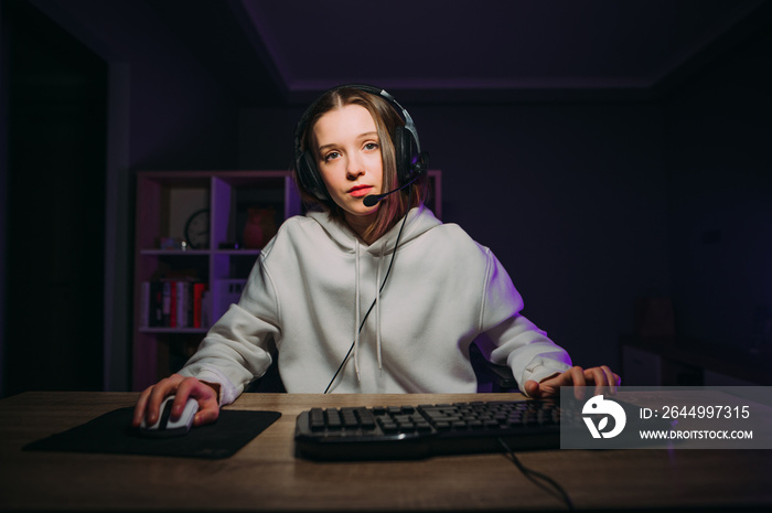 Attractive female gamer in a headset on her head plays games at home at night at the table and streams, looking focused at the camera.