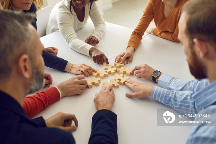 Happy team of multiethnic business people join puzzle pieces sitting around office table. Group of multiracial men and women put jigsaw parts together as metaphor for teamwork and looking for solution