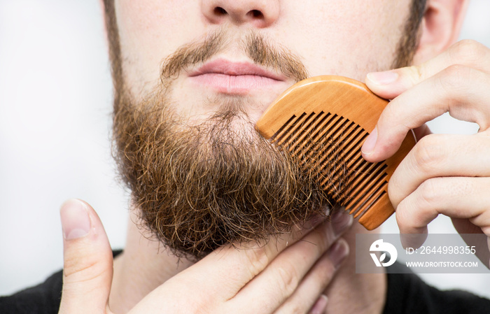 Closeup of a young man styling his long beard with a comb while standing alone in a studio against a white background