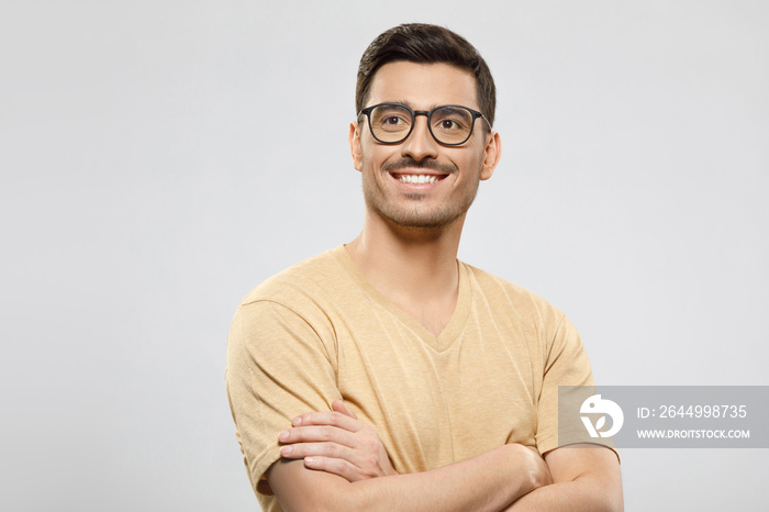 Portrait of young handsome guy dressed in beige t-shirt and wearing eyeglasses, holding arms crossed, looking aside with smile as if thinking, isolated on gray background