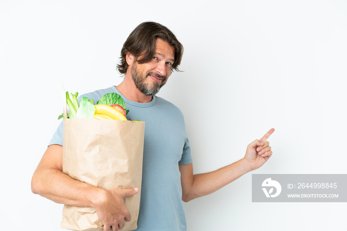 Senior dutch man holding grocery shopping bag over isolated background pointing back