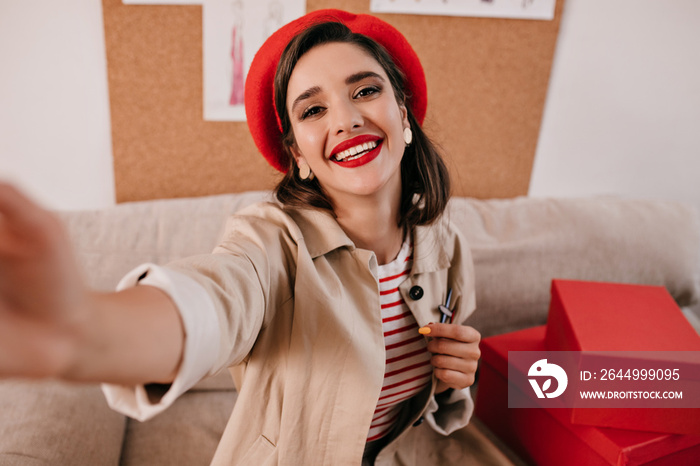 Brunette woman in red beret and trendy trench takes selfie. Smiling young lady with bright lips in red hat and a striped sweater makes photo..