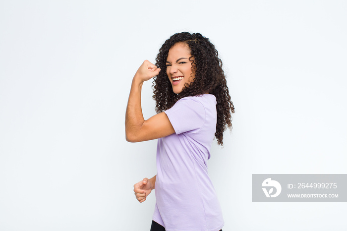 young black woman feeling happy, satisfied and powerful, flexing fit and muscular biceps, looking strong after the gym against white wall