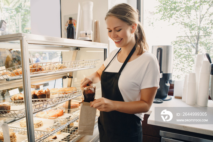 Portrait of white Caucasian beautiful barista woman taking muffin pastry from shop-window. Person at work place small business concept