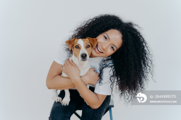 Happy curly woman tilts head, holds pedigree dog, has cheerful expression, smiles pleasantly, has curly hairstyle, wears white t shirt, isolated. Afro lady petting favourite domestic animal.