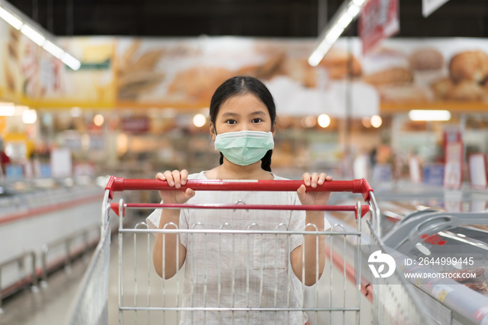 Asian child or kid girl wearing face mask for close mouth nose and flu cough sick to protect covid-19 or dust with pushing supermarket trolley or cart for shopping in market mall or department store