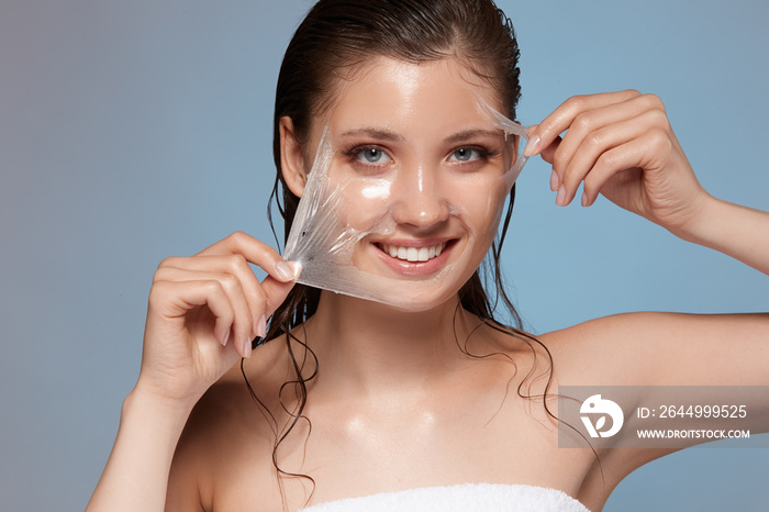 young and happy female peeling off facial mask and smiling to the camera, woman treats her face in bath towel