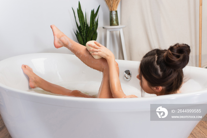 young woman with hair bun taking bath with with loofah in bathtub