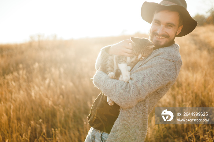 Lovely young hipster with a cat. A guy with a mustache and a beautiful smile is hugging a cat. Beautiful autumn sunset.