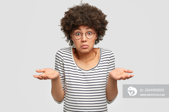 Uncertain African American female with dark skin, shruggs shoulders and asks something, has clueless expression, wears glasses and striped sweater, isolated on white background. Woman faces dilemma