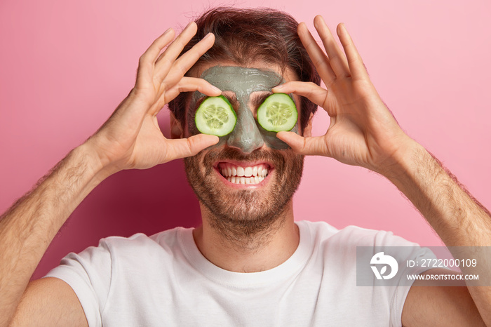 Beauty portrait of cheerful man poses with clay mask on face, two slices of cucumber on eyes, wears 
