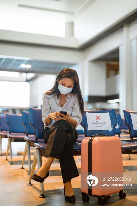 戴着口罩的年轻亚洲旅游女性在机场打电话，坐在远处