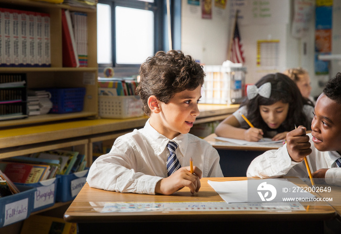 Portrait of two smiling schoolboys (8-9 and 10-11) sitting at adjacent desks in classroom talking